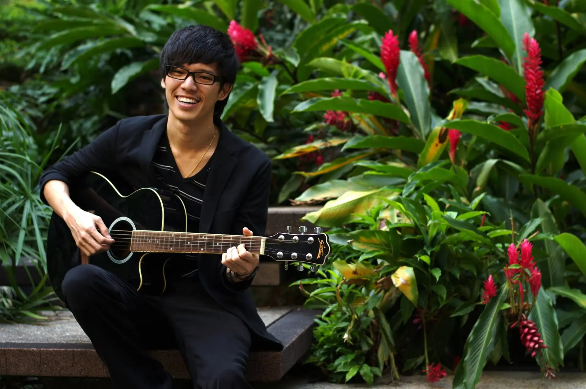 Dr. Jovian Tan smiling as he plays an acoustic guitar, seated on garden steps among tropical plants and red ginger flowers