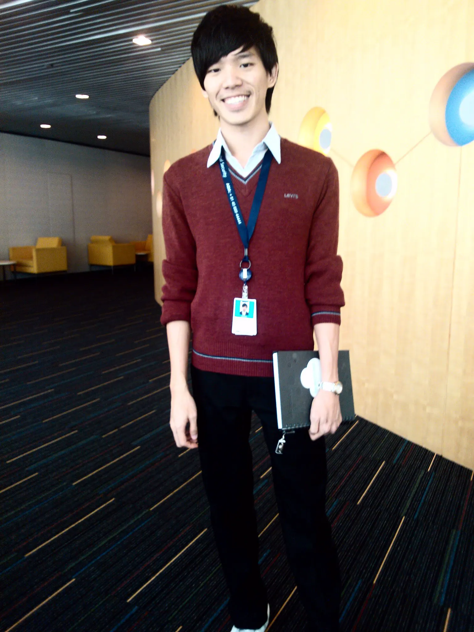 Dr. Jovian Tan smiling in a red sweater with a staff lanyard and notebook, standing in a corridor at a Microsoft office