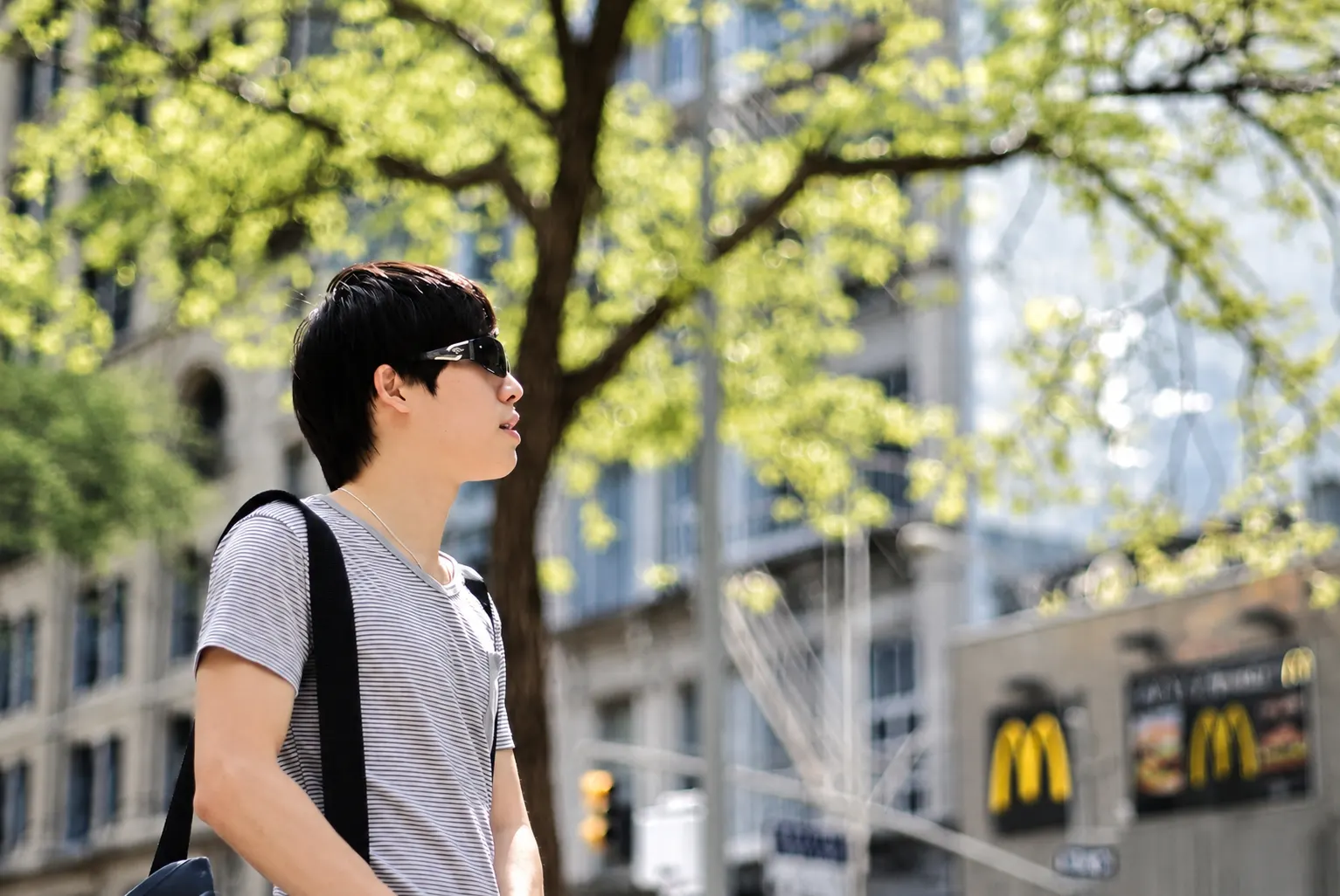 Dr. Jovian Tan in sunglasses and a striped T-shirt, carrying a shoulder bag and looking up beneath a tree among city buildings