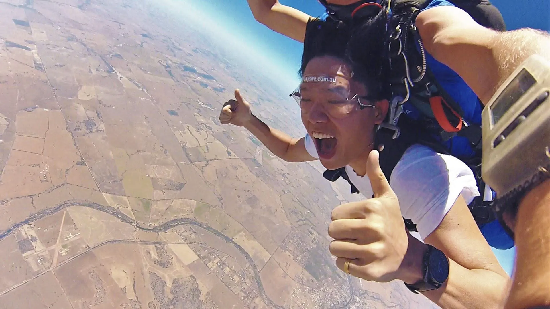 Dr. Jovian Tan in freefall during a tandem skydive, grinning with both thumbs up, farmland and a river far below