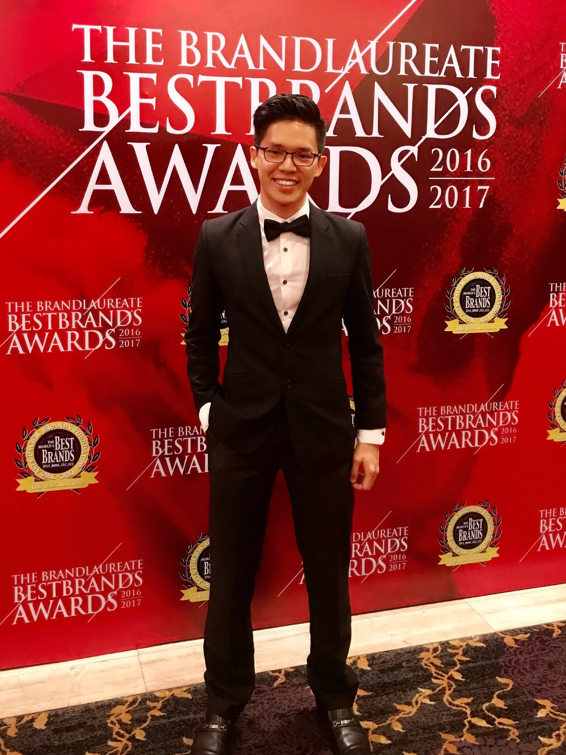 Dr. Jovian Tan in a black tuxedo and bow tie in front of The BrandLaureate BestBrands Awards 2016–2017 backdrop
