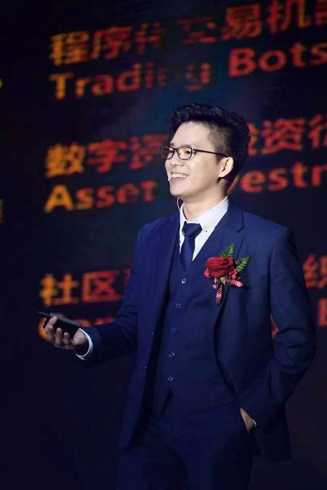 Dr. Jovian Tan in a navy three-piece suit with a rose boutonnière, presenting on stage in front of a screen listing trading bots and asset investment