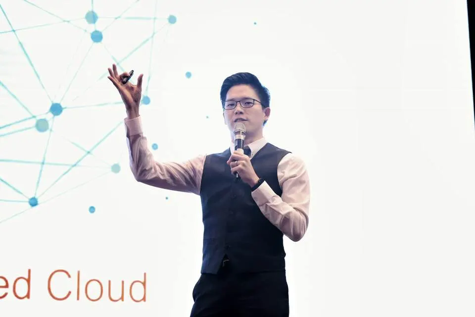 Dr. Jovian Tan on stage with a microphone, gesturing towards a slide showing a network diagram