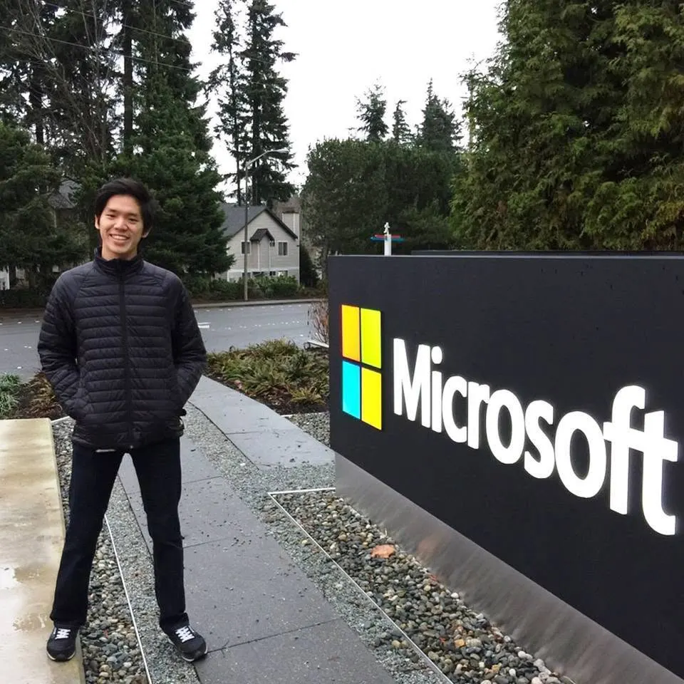 Dr. Jovian Tan in a black puffer jacket, smiling beside a large Microsoft logo sign on a tree-lined campus