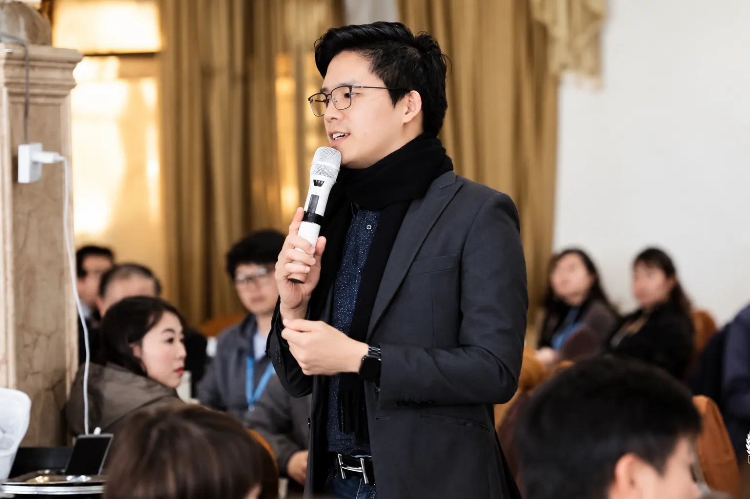 Dr. Jovian Tan in a dark blazer and scarf speaking into a microphone, standing among a seated conference audience