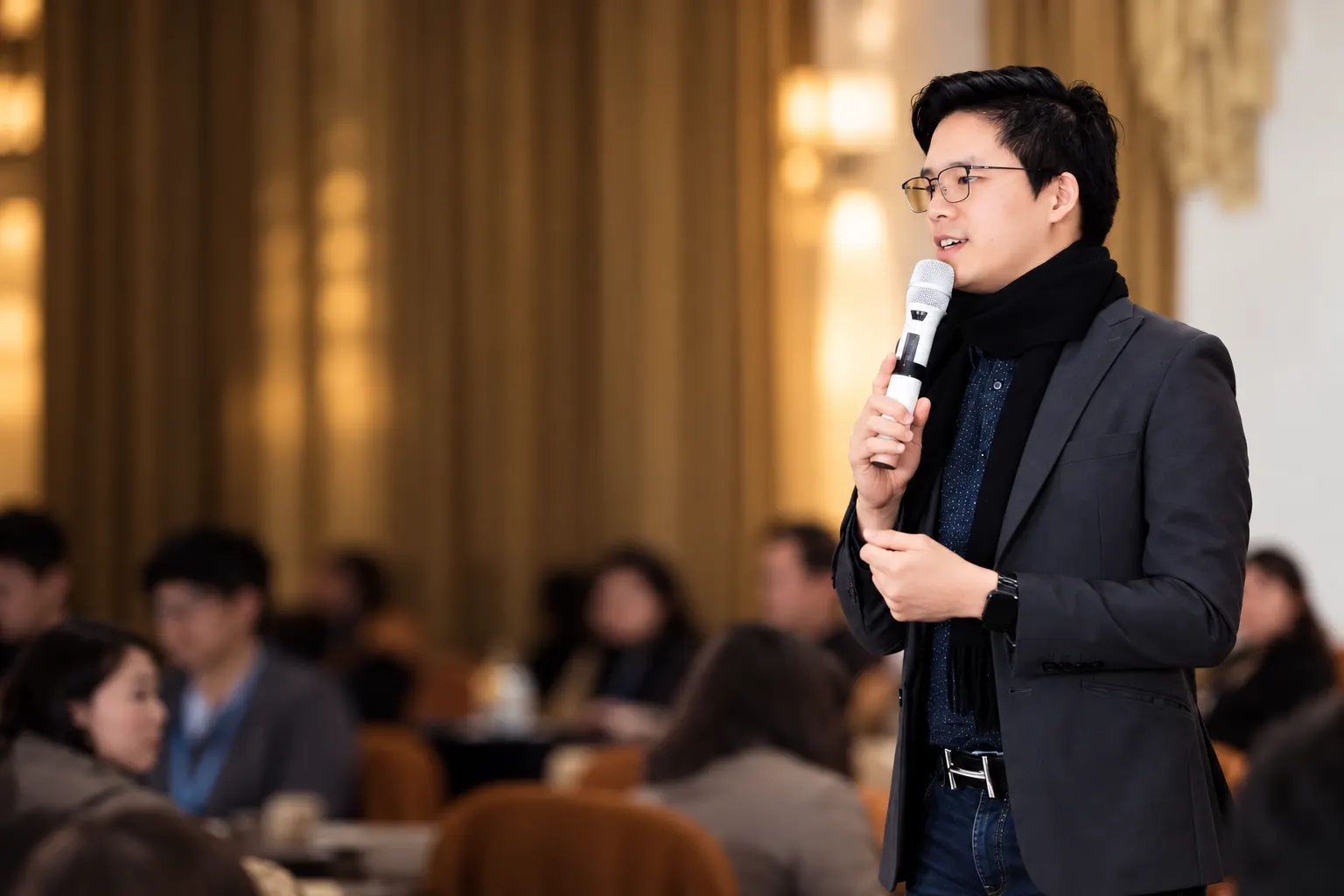 Dr. Jovian Tan in a dark blazer and black scarf speaking into a microphone at a conference, with a seated audience and warm golden curtains behind him